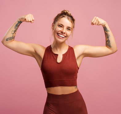 Smiling fit woman showing her muscles on hands, looking at camera, isolated on pink